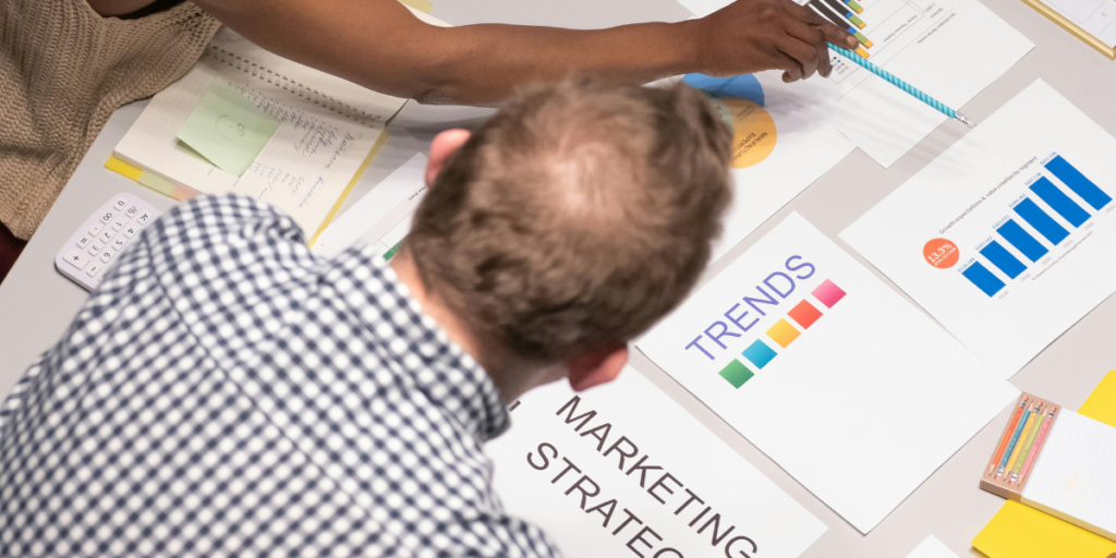 An overhead view of a person in a checkered shirt examining various marketing materials, including graphs and charts, with a calculator and papers on the table indicating an in-depth strategic planning session